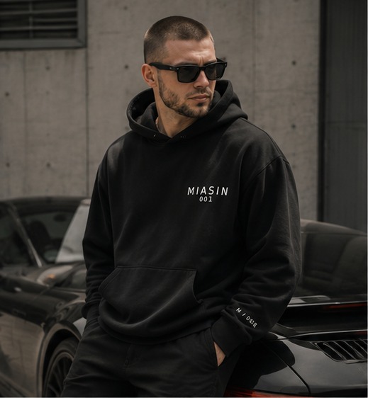 A model wearing the black MIASIN 001 hoodie beside a black car