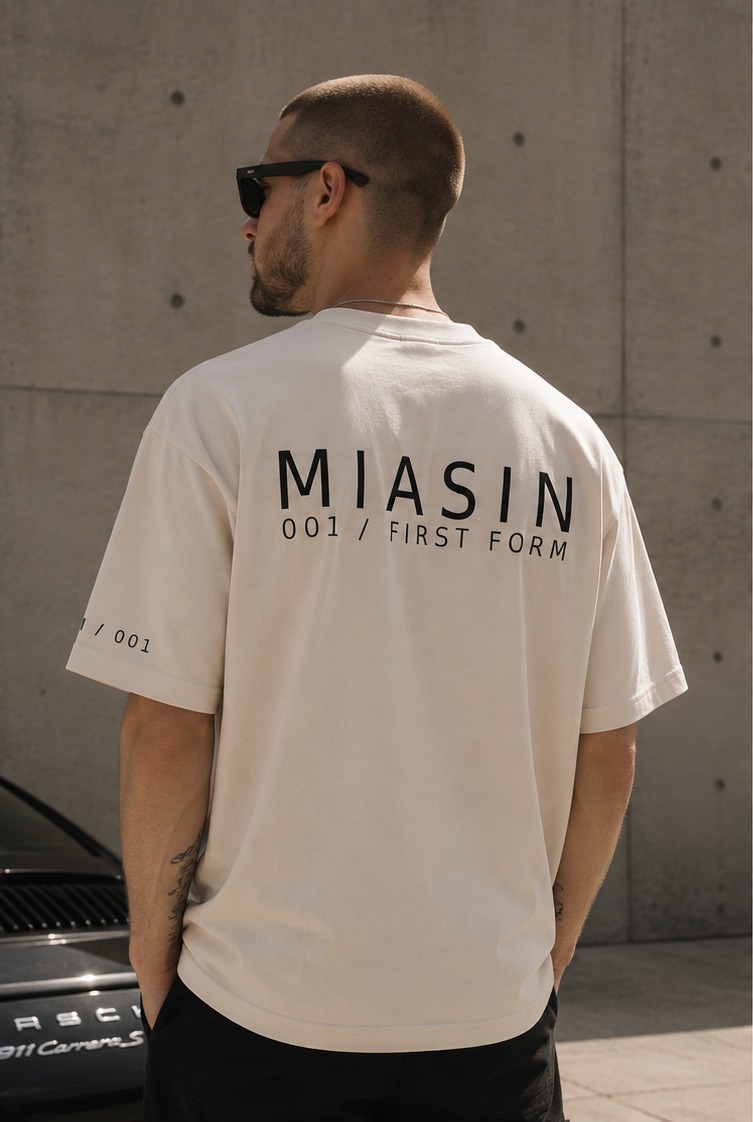 A model photographed from behind wearing the bone MIASIN 001 Box Tee against a concrete wall