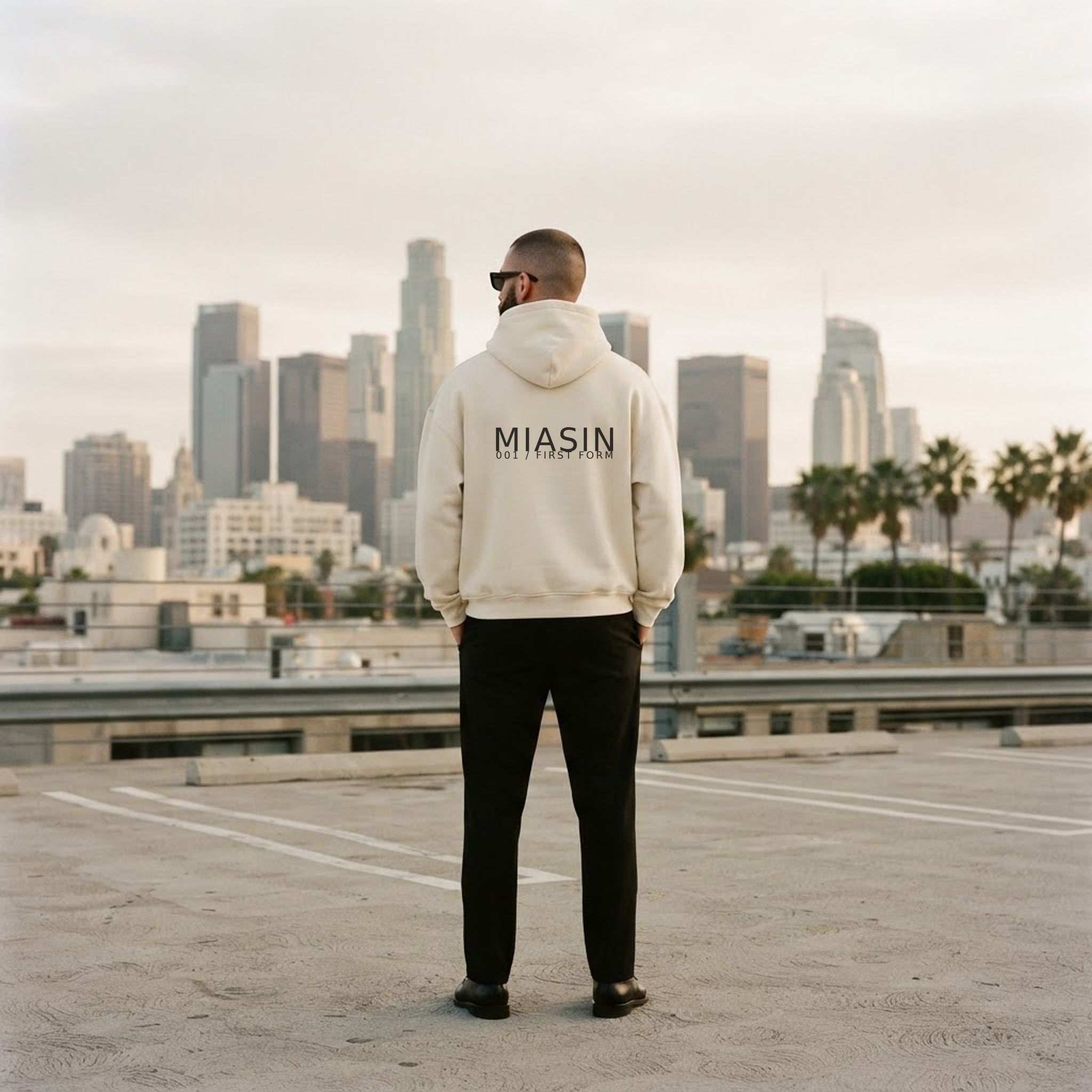 A model photographed from behind wearing the bone MIASIN 001 hoodie on a rooftop above the downtown Los Angeles skyline